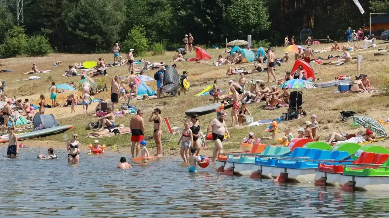 Tłumy ludzi wypoczywają na plaży nad jeziorem Chechło. Woda jest pełna kąpiących się, a na brzegu opalają się plażowicze.