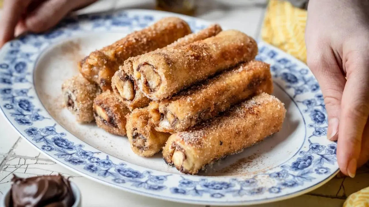 Nutella French Toast Rolls, goldbraun gebacken und mit Zimt-Zucker bestreut, auf einem Teller mit blauem Blumenmuster.