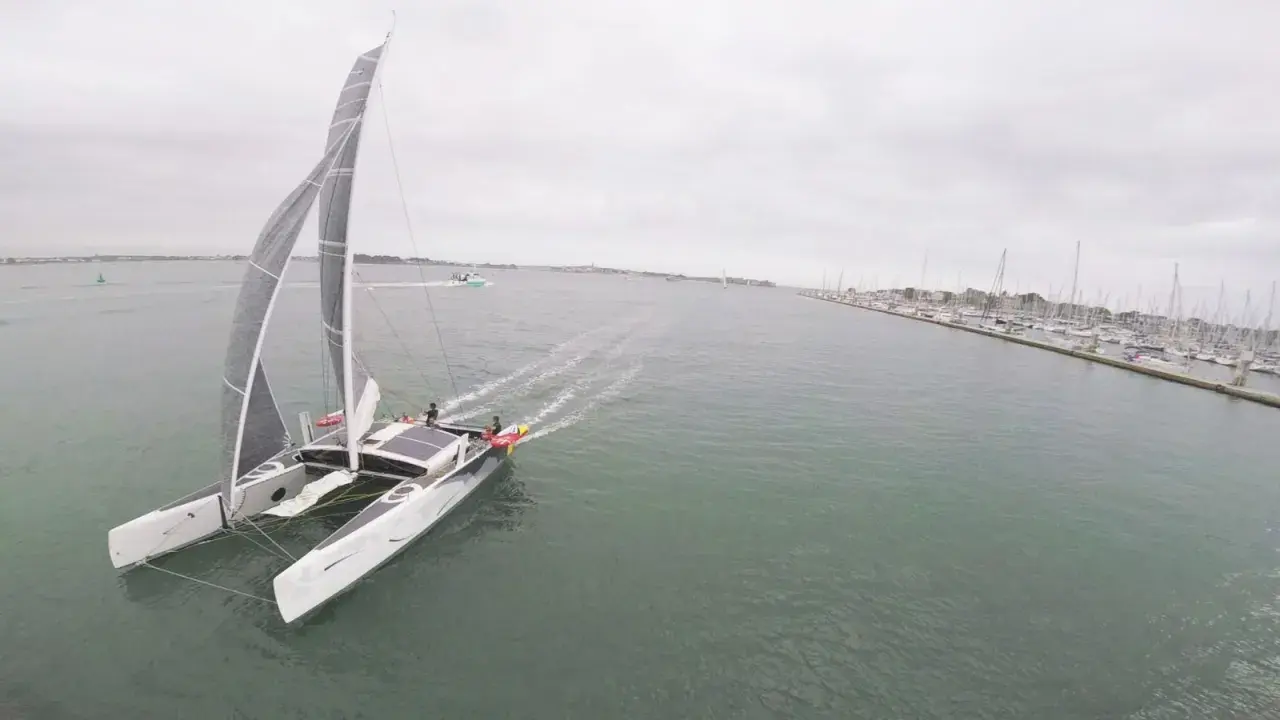 Un catamaran TS3 navigue sur une mer calme, avec un port rempli de voiliers à droite.