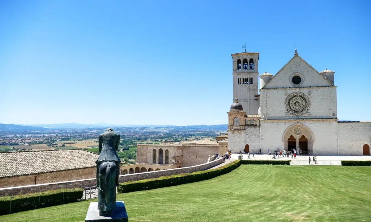 Statua equestre domina il prato verde di Assisi. Cosa vedere: la Basilica di San Francesco, con la sua imponente facciata e campanile, si staglia contro il cielo azzurro.
