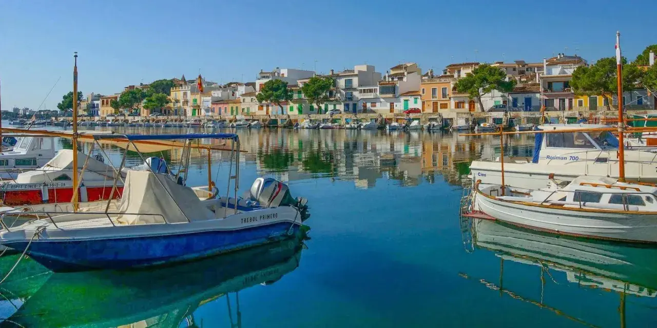 Boote im Hafen von Portocolom, Mallorca, spiegeln sich im ruhigen, blauen Wasser vor bunten Häusern.
