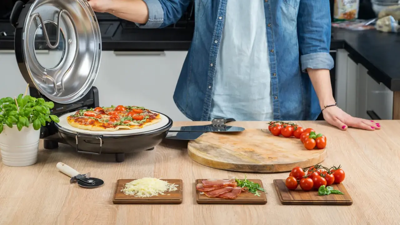 Otwarta opiekacz do pizzy, na kamieniu piec pizza z pomidorkami i bazylią. Obok deska z pomidorkami, startym serem i szynką.