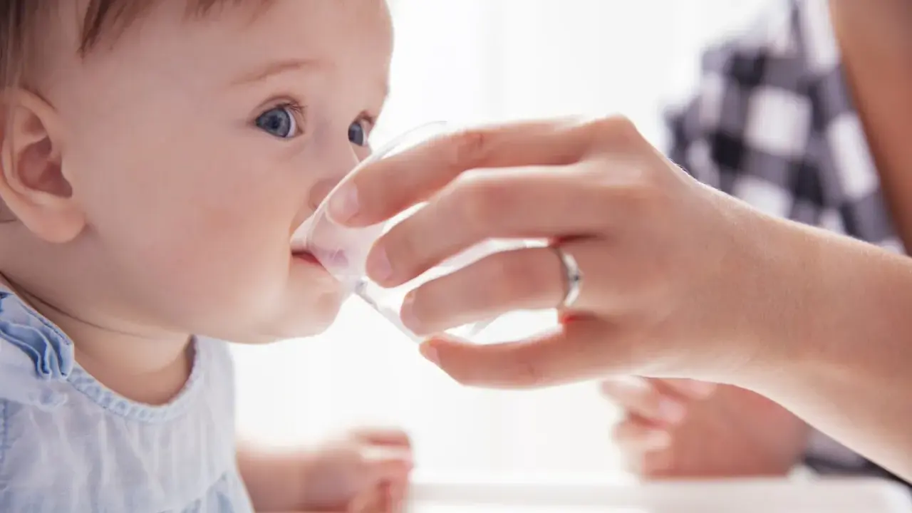 Ein Baby trinkt vorsichtig Wasser aus einem Becher, gehalten von einer Hand. Die Frage "ab wann Baby Wasser geben?" wird hier visuell beantwortet.