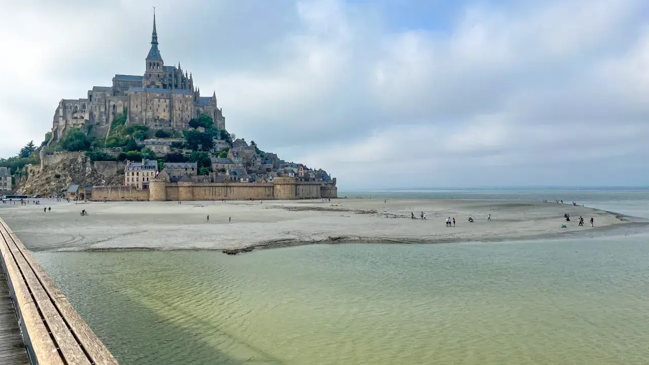 St. Michel, eine beeindruckende Abtei auf einer Felseninsel, ragt über einer sandigen Bucht auf. Menschen spazieren am Strand, während das Wasser sanft ansteigt.