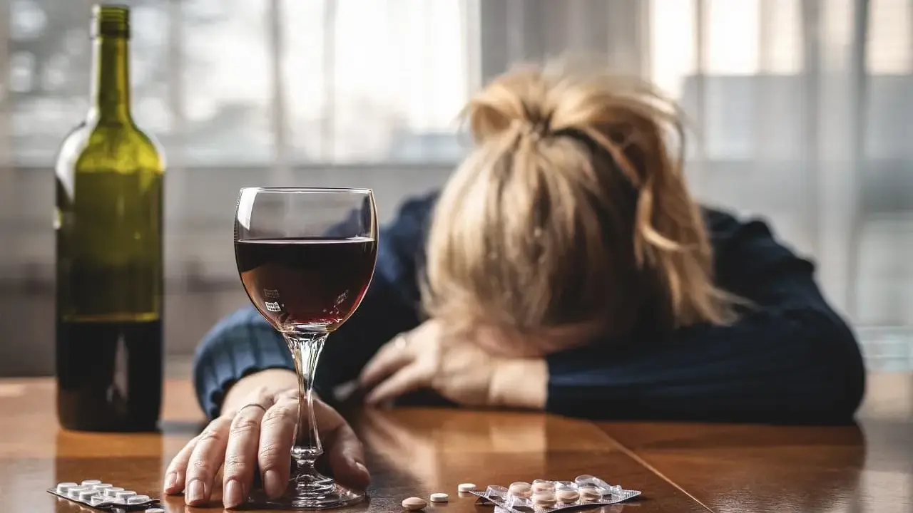 Kobieta z głową opartą na rękach, obok kieliszek wina, butelka i opakowania psychotropów.