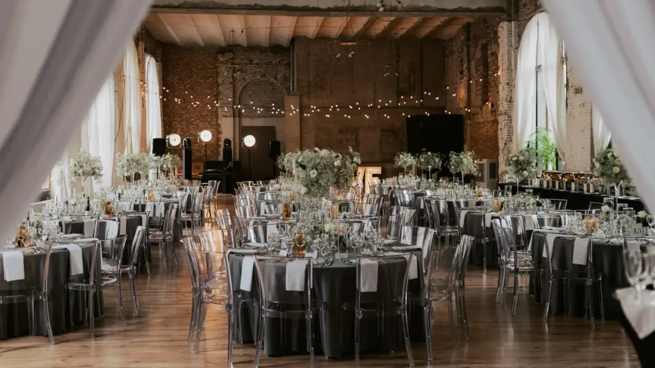 Eleganckie wesele w industrialnym stylu. Stoły nakryte szarymi obrusami, ozdobione białymi kwiatami i transparentnymi krzesłami. Girlandy świateł tworzą magiczny nastrój.
