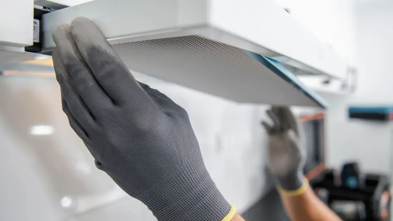 Hände mit Handschuhen ziehen einen Filter aus einer Dunstabzugshaube, um den Motor zu reinigen.