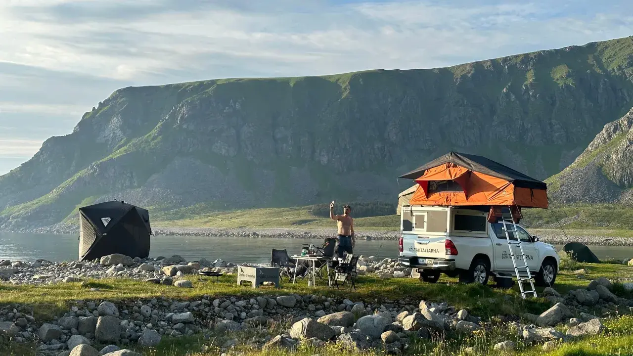 En man tar en selfie vid sitt tak-tält på en strand i Norge. Bredvid står en mörk kupoltält och ett bord med stolar. Berg i bakgrunden.