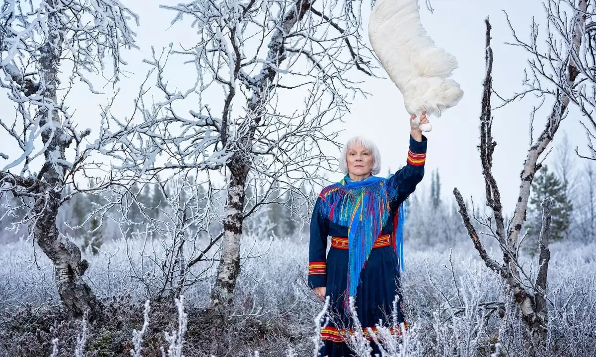 Britta Marakatt Labba i samedräkt håller upp en stor vit fjäder i en snötäckt skog.