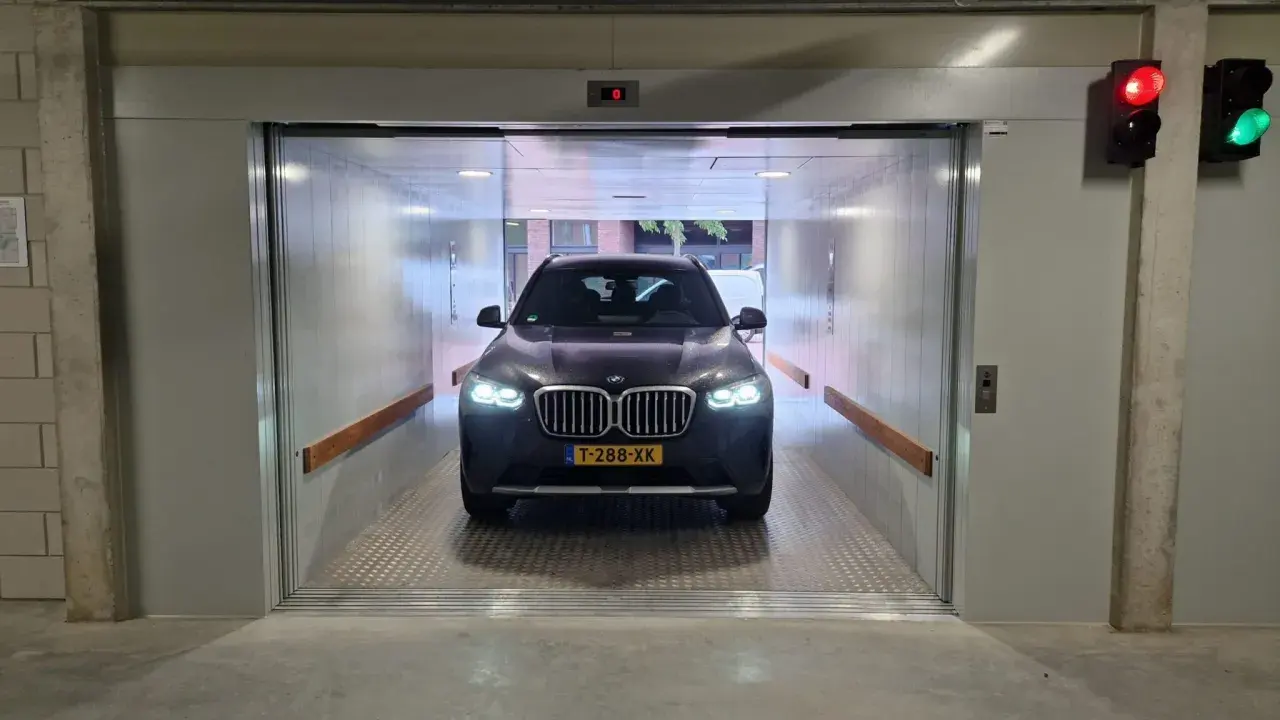 Un coche BMW oscuro espera en un montacoches para garaje. La luz roja del semáforo indica que debe esperar.