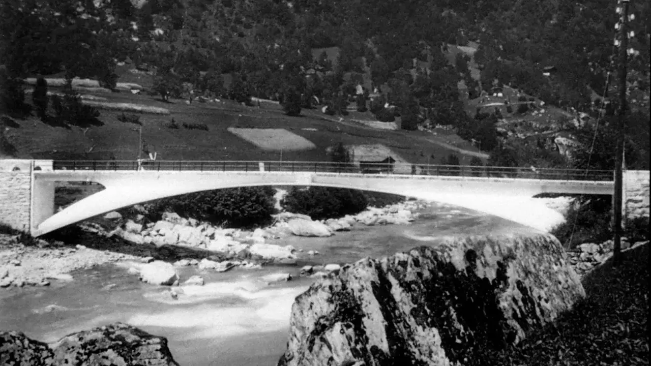 Puente de arco de hormigón de **Robert Maillart** sobre un río rocoso, con colinas boscosas al fondo.