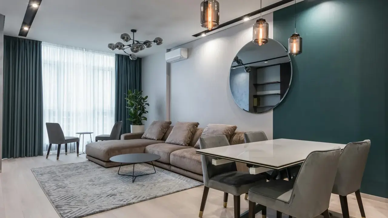 Nowoczesna lampa do salonu, wisząca nad stołem, tworzy przytulną atmosferę. W tle duża sofa i okno z zasłonami.