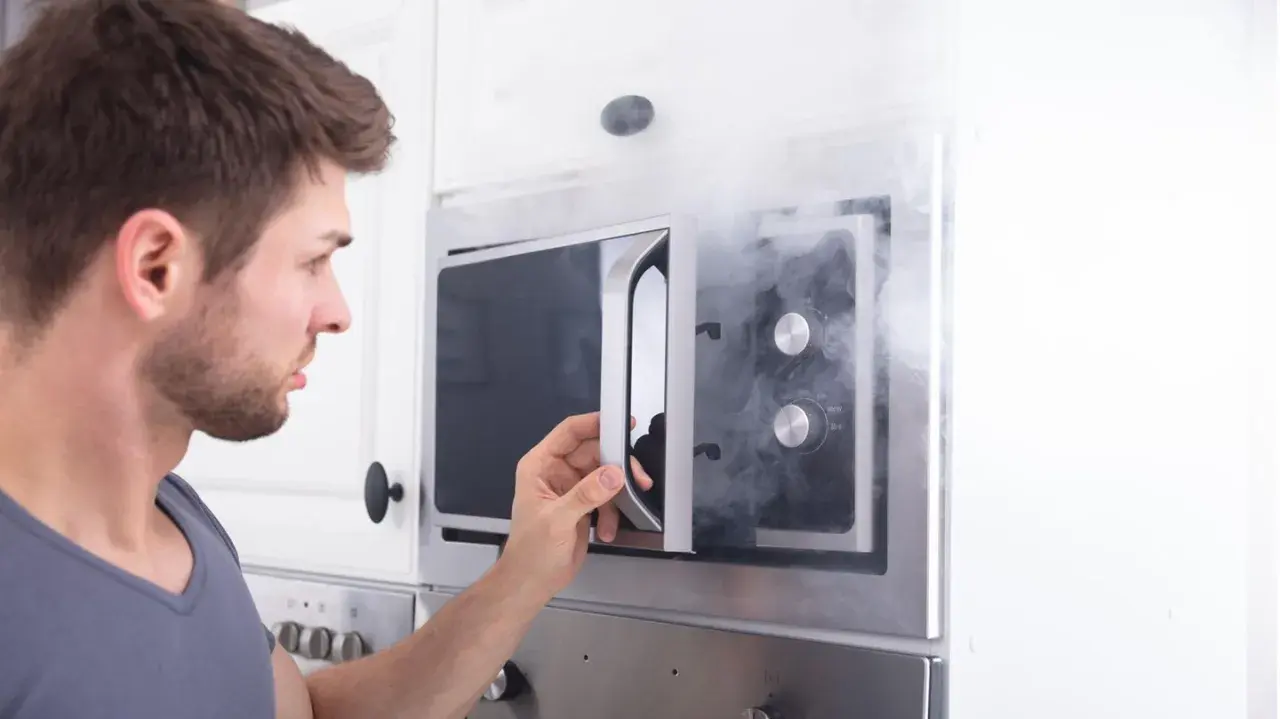 Mann öffnet Mikrowelle, aus der Rauch dringt. Ein Fehler mit Metall in Mikrowelle kann gefährlich sein.