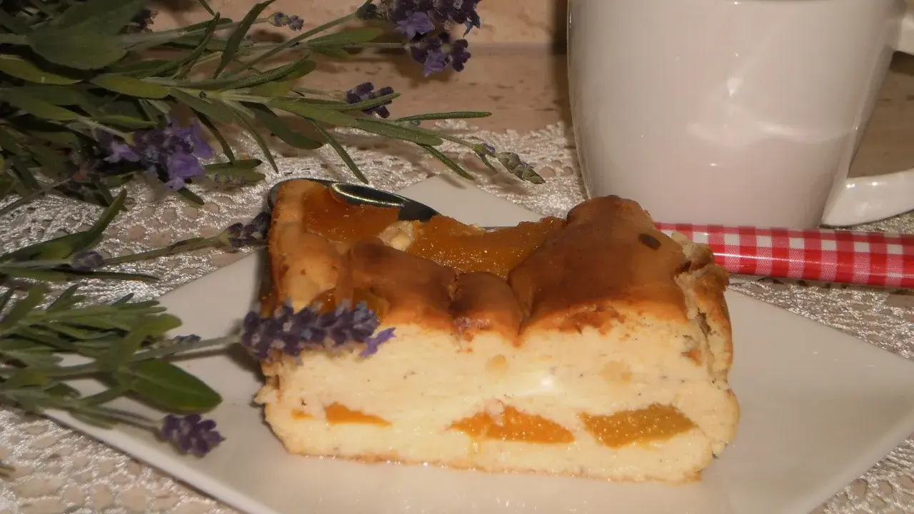 Pyszny sernik z brzoskwiniami, udekorowany gałązką lawendy, obok białej filiżanki i czerwono-białej łyżeczki.