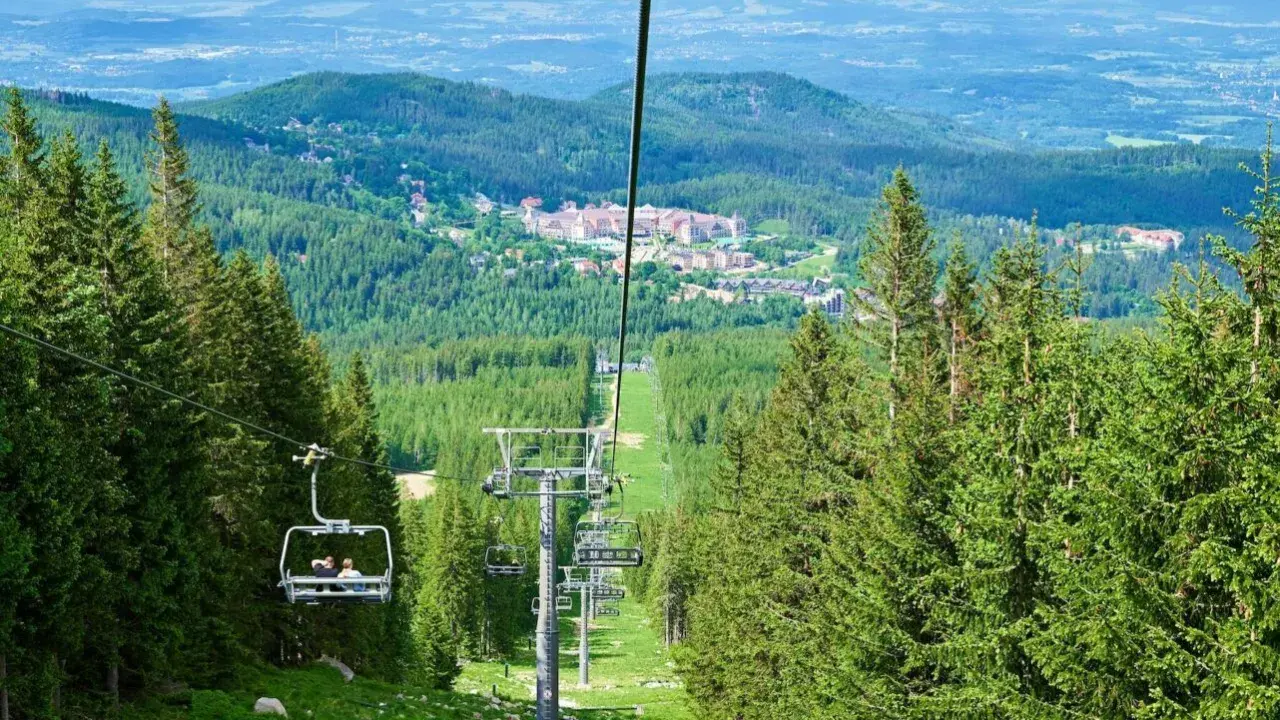 Wjazd kolejką na wysoką kopę trasę, otoczoną lasem świerkowym. W oddali widać zabudowania i góry.