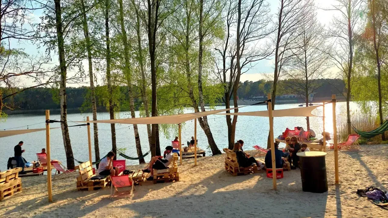 Pola namiotowe nad jeziorem, ludzie odpoczywają na plaży pod baldachimami, wśród drzew.