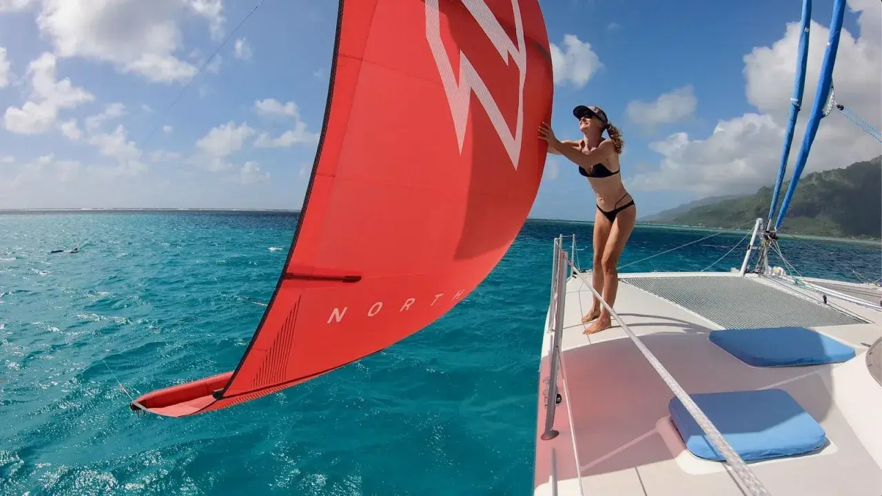 Une femme en bikini ajuste la voile rouge d'un petit catamaran habitable. L'eau turquoise et le ciel bleu créent un décor paradisiaque.