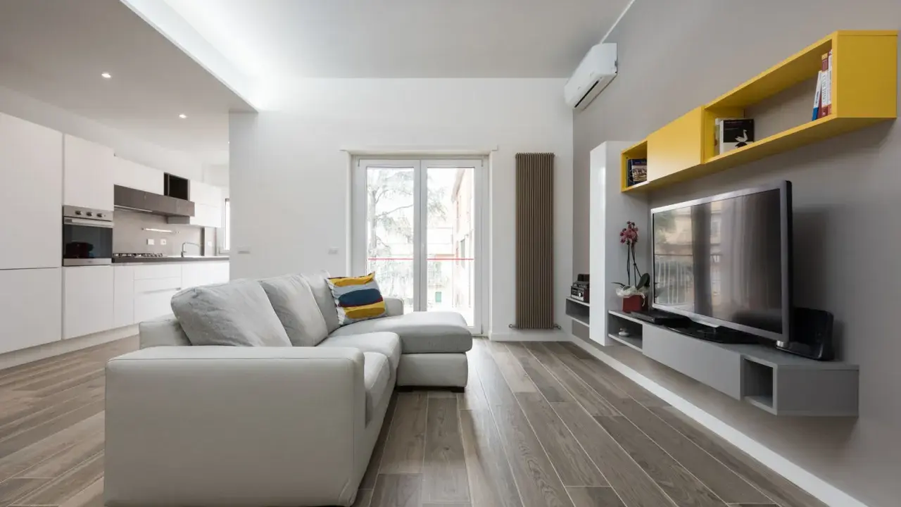 Un progetto soggiorno stretto e lungo con divano grigio, TV a parete e mensole gialle. La cucina moderna è visibile sullo sfondo.