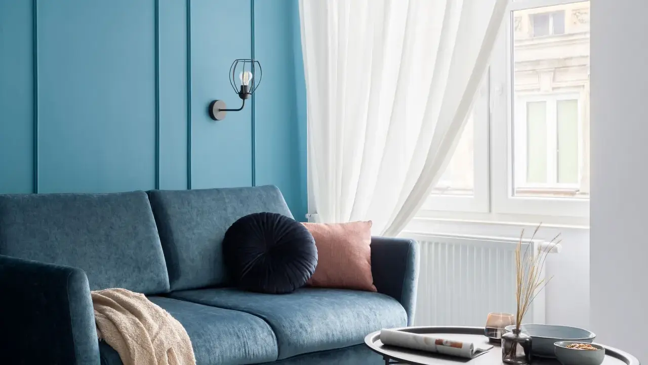 Divano blu petrolio con cuscini decorativi e coperta. Parete azzurra con applique. Abbinamenti moderni e rilassanti.
