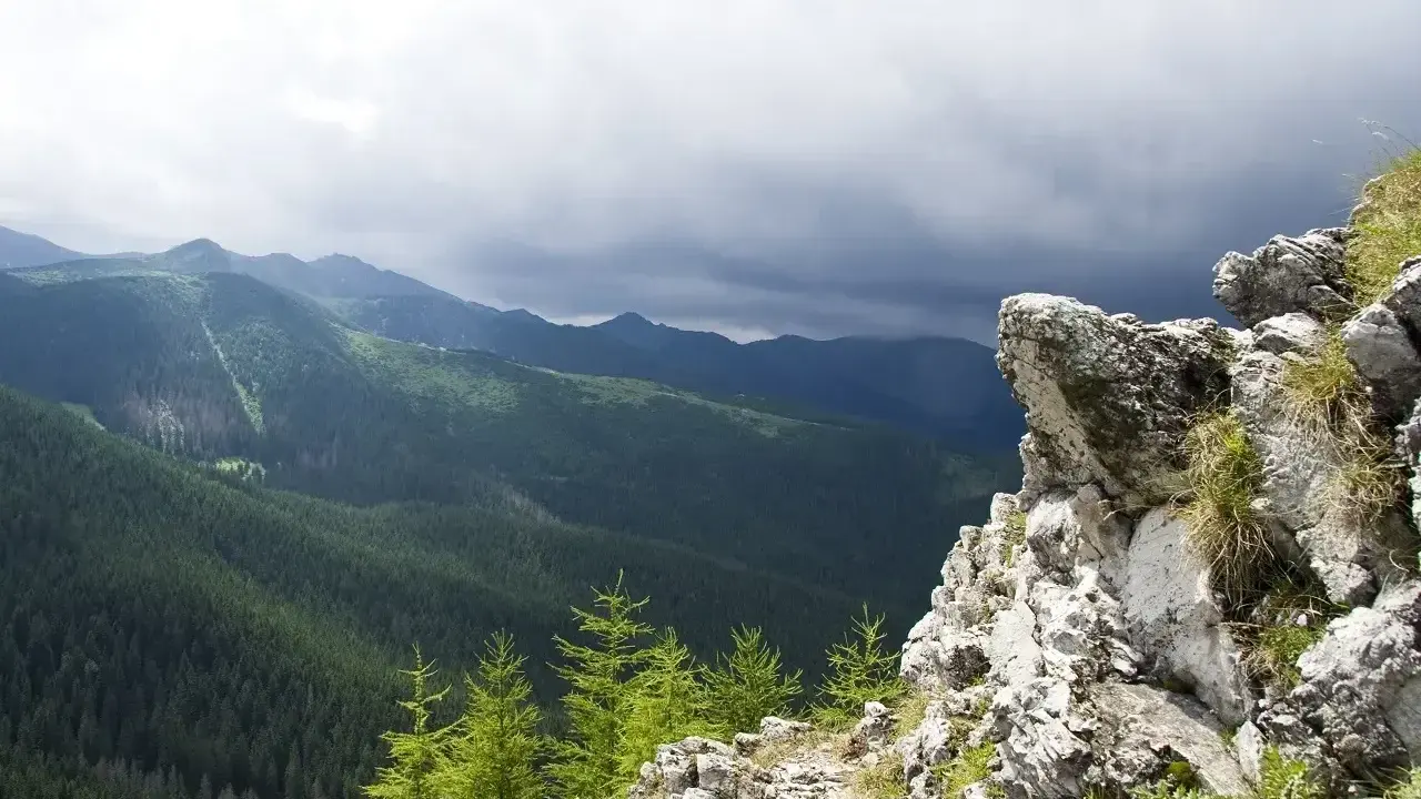 Skalisty szczyt w Zakopanem z widokiem na gęsto zalesione doliny Tatr pod burzowym niebem.
