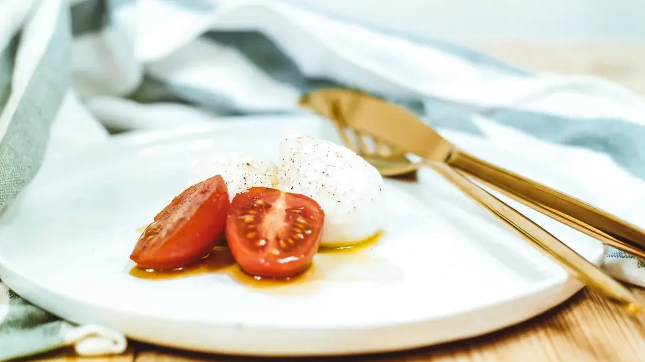 Kuleczki mozzarelli z pomidorkami koktajlowymi na białym talerzu, obok złote sztućce i ściereczka.
