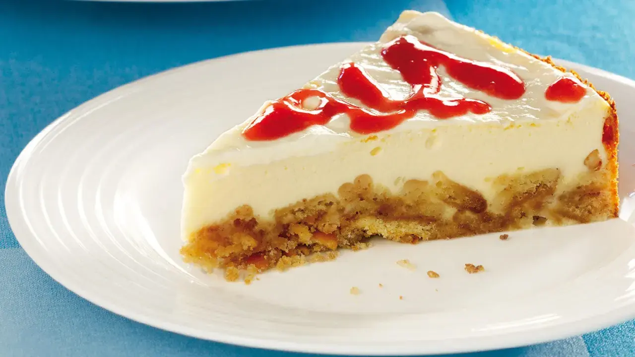 Ein Stück cremiger cheesecake mit keksboden, garniert mit rotem Fruchtpüree auf einem weißen Teller.