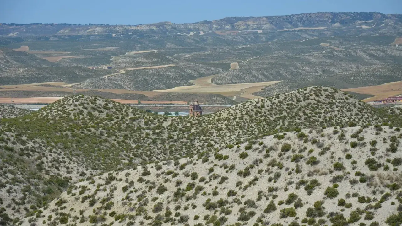 Paisaje árido del desierto de Monegros con colinas cubiertas de matorral y una torre de iglesia solitaria al fondo.