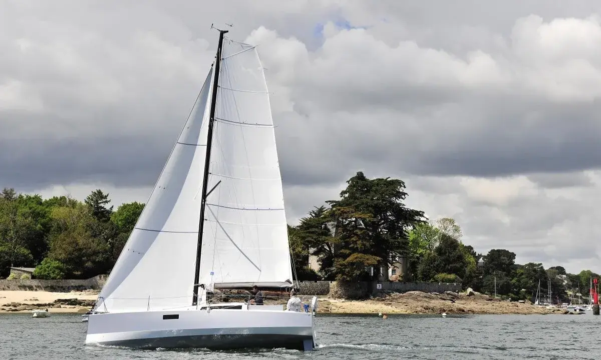 Voilier blanc moderne naviguant sur l'eau, avec des avis sur le Pogo 30. Paysage côtier verdoyant en arrière-plan.