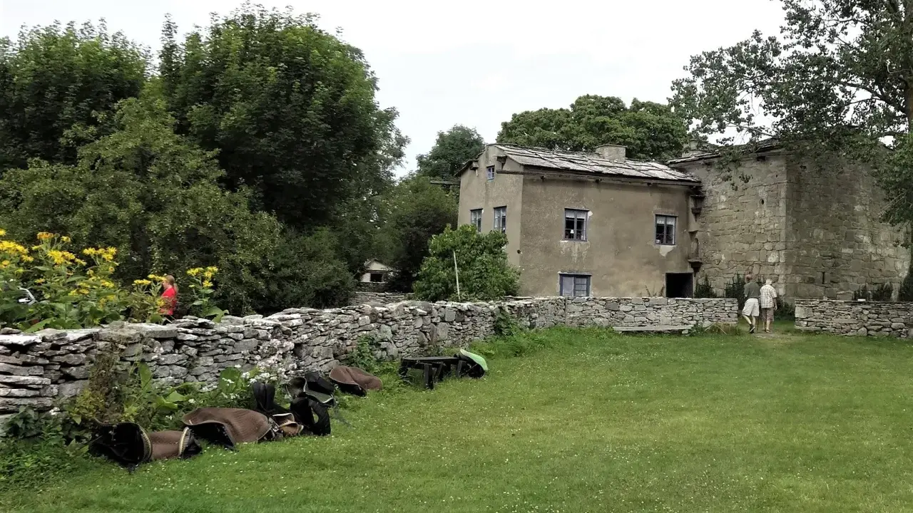 En gammal byggnad med stenväggar och ett tak av skiffer. En person i rött går förbi gula blommor vid en stenmur. Kanske en framtida vingård på Gotland?