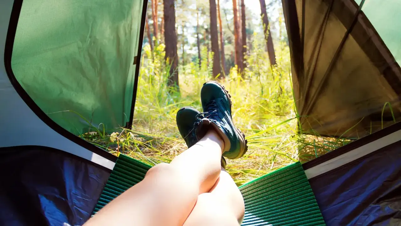 Nogi w butach trekkingowych odpoczywają w namiocie. Słońce przebija się przez drzewa. Idealne miejsce, by zanocuj w lesie.
