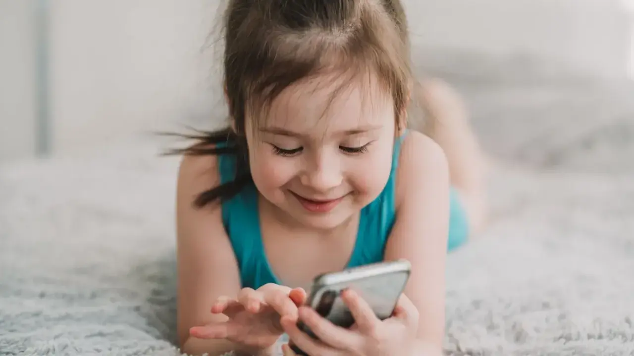 Uśmiechnięta dziewczynka leży na dywanie, bawiąc się telefonem. Wpływ telefonu komórkowego na mózg dziecka jest tematem dyskusji.