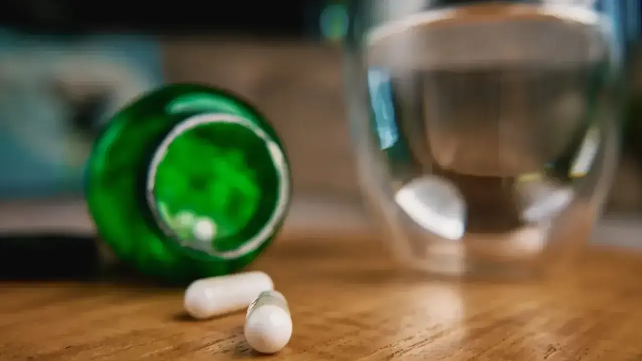 Berberyna a alkohol: dwie białe kapsułki obok zielonego opakowania i szklanki z wodą.