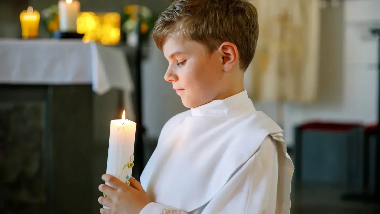 Chłopiec w białej albie trzyma zapaloną świecę podczas uroczystości komunii świętej.