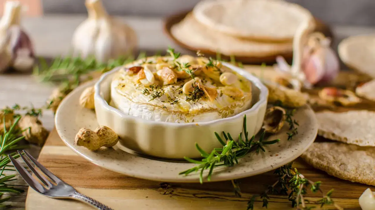 Pieczony camembert w naczyniu, ozdobiony ziołami i orzechami, z czosnkiem i pieczywem w tle.