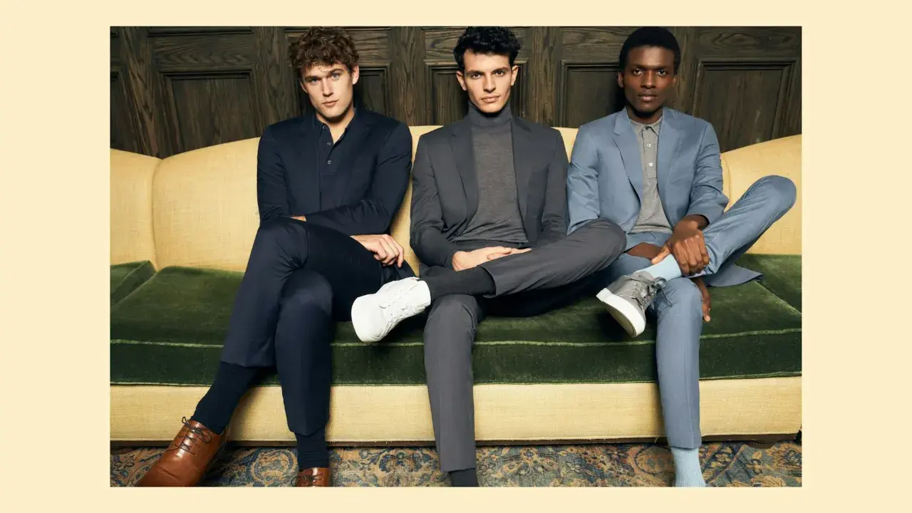 Three men in suits sit on a couch. The man on the left wears dark dress pants, while the other two wear suit pants.