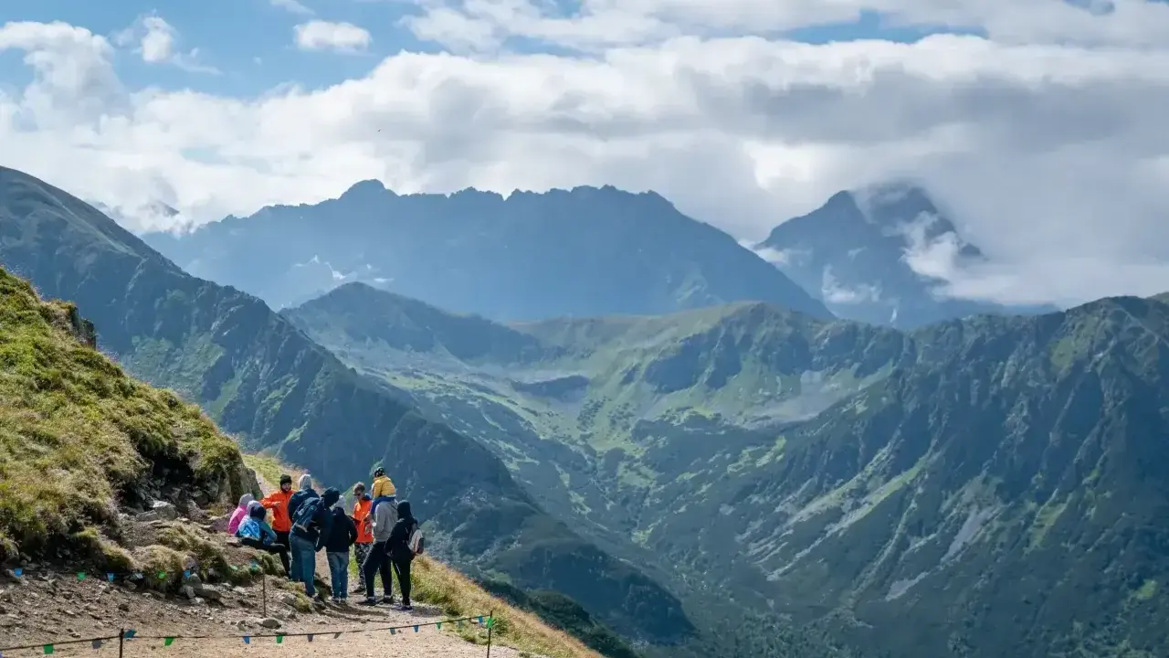 Grupa turystów odpoczywa na szlaku z widokiem na majestatyczne Tatry. W oddali majaczy Kasprowy Wierch, otoczony chmurami.