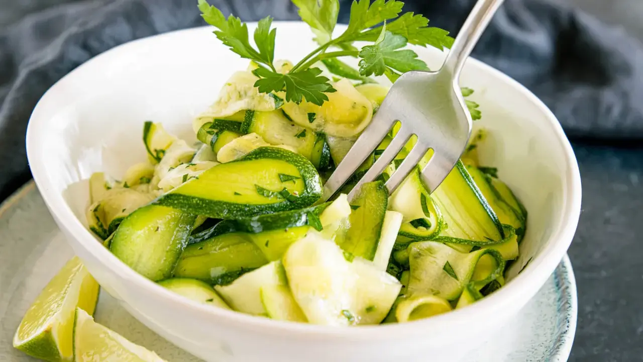 Frischer Zucchini-Salat mit Petersilie und Limettenspalten, serviert in einer weißen Schüssel. Eine Gabel steckt im Salat.