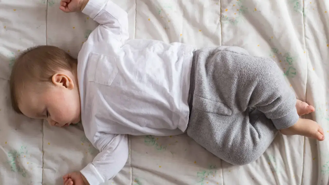 Un neonato non dorme, ma giace sdraiato su una coperta con un pigiamino bianco e grigio.