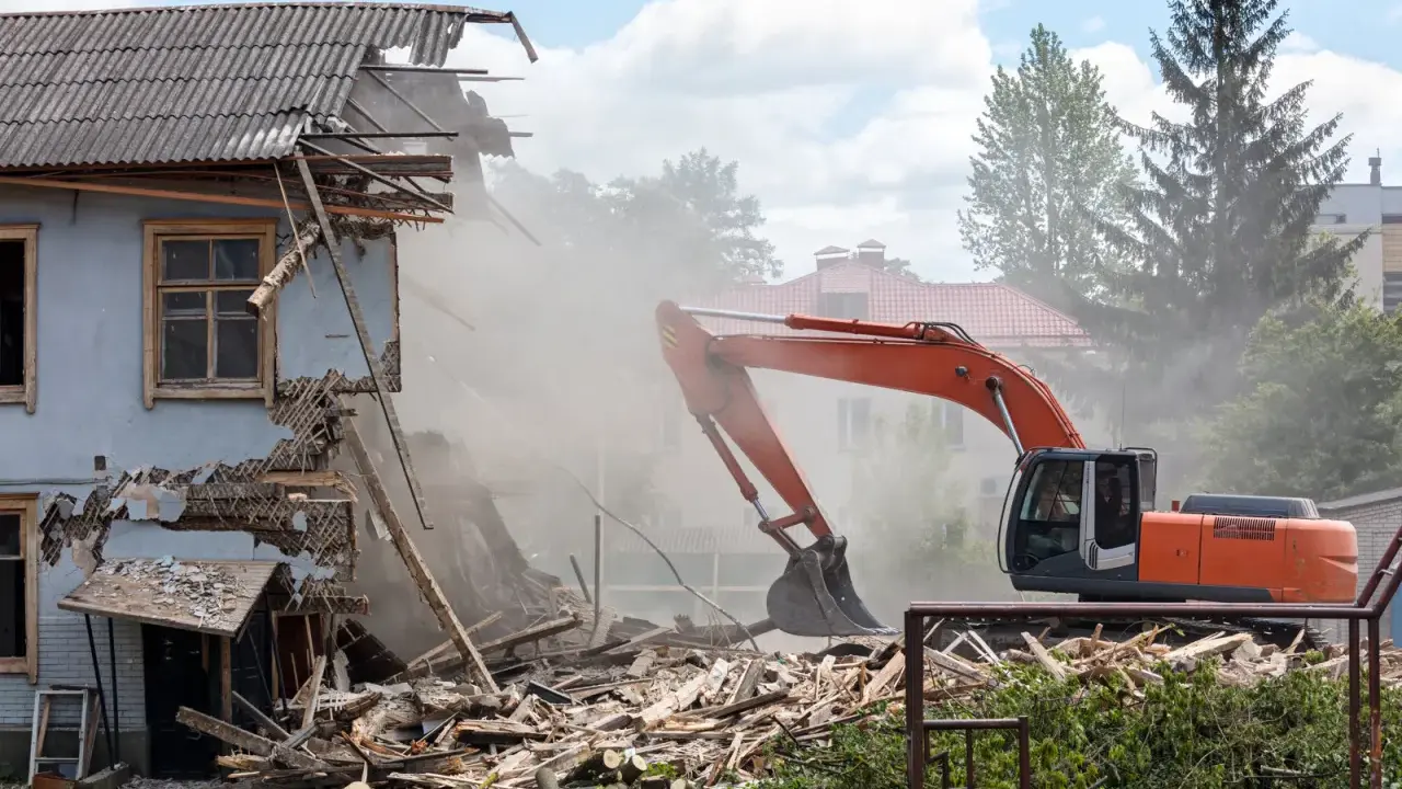 Pomarańczowy koparka prowadzi rozbiórkę budynku gospodarczego, wzbijając tumany kurzu. Ruiny i drewno leżą na ziemi.