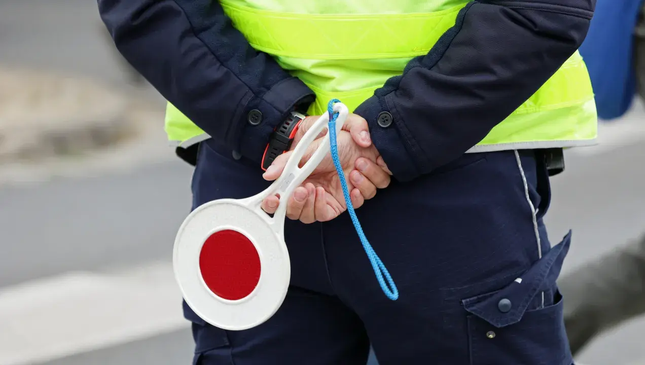 Policjant w kamizelce odblaskowej trzyma za plecami lizak do zatrzymywania pojazdów.