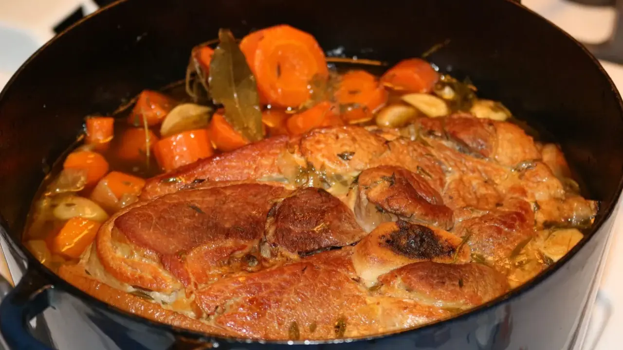 Rouelle de porc mijotant dans une cocotte avec des carottes, de l'ail et des herbes.