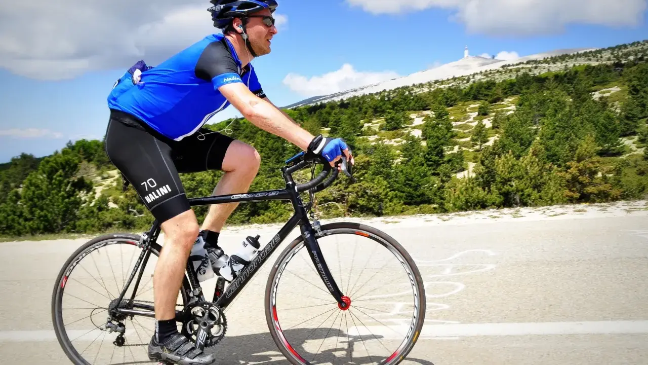 Ciclista in maglia blu affronta la **salita** del **Mont Ventoux**, con la cima innevata in lontananza.