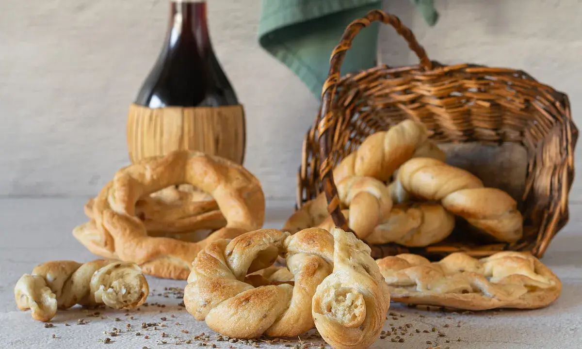 Pane intrecciato e ciambelle, ispirazione per ricette ciociare, accanto a una bottiglia di vino rosso e un cesto di vimini.