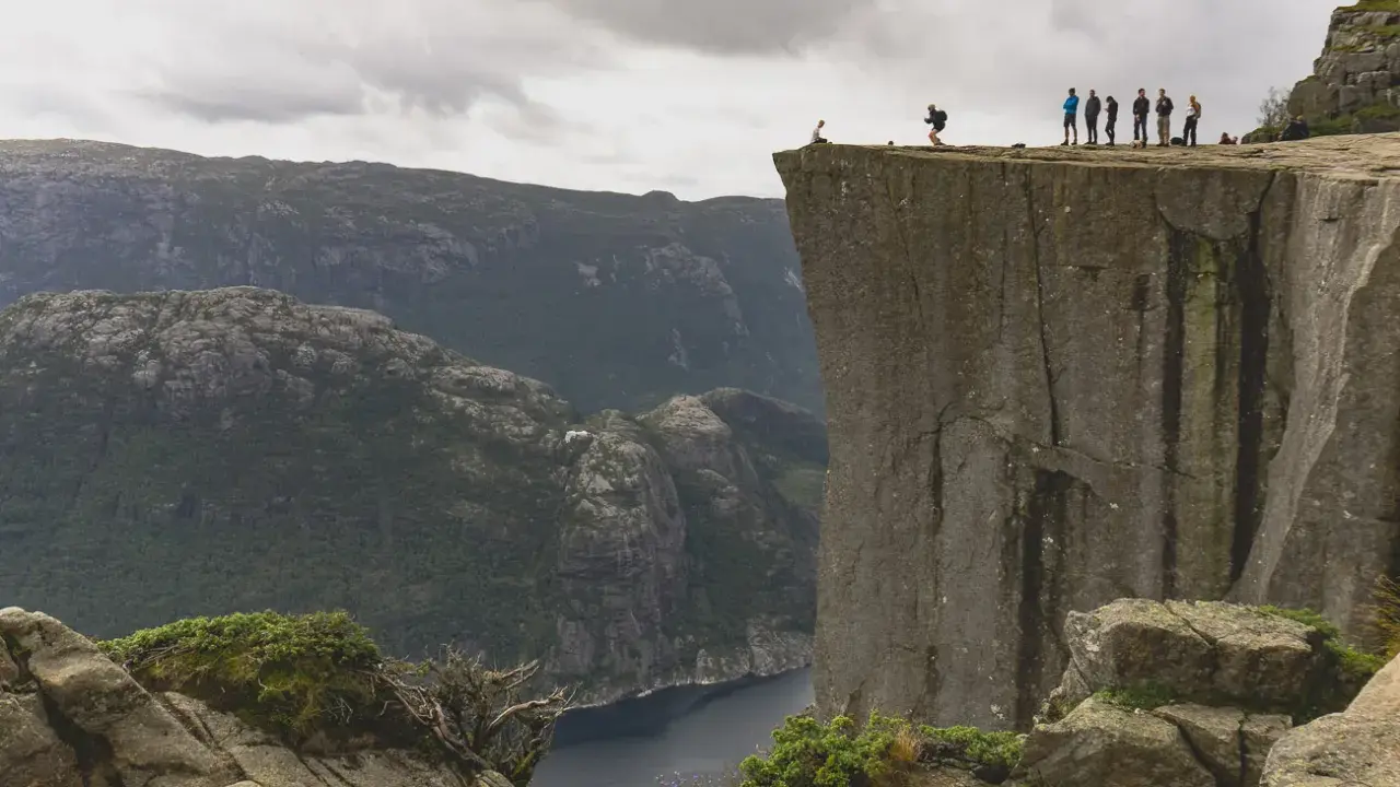 Grupa ludzi na krawędzi klifu w **Stavanger, Norwegia**, z widokiem na fiord i góry.