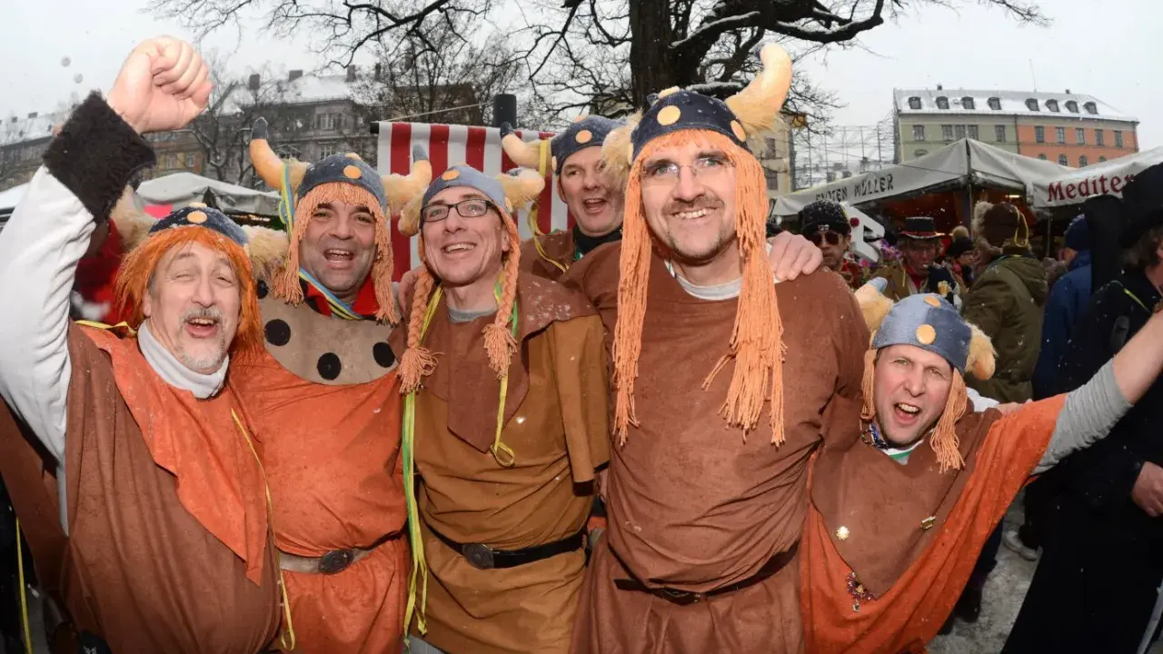 Fröhliche Männer in historischer Wikinger-Kleidung mit Hörnerhelmen und orangefarbenen Perücken feiern ausgelassen.