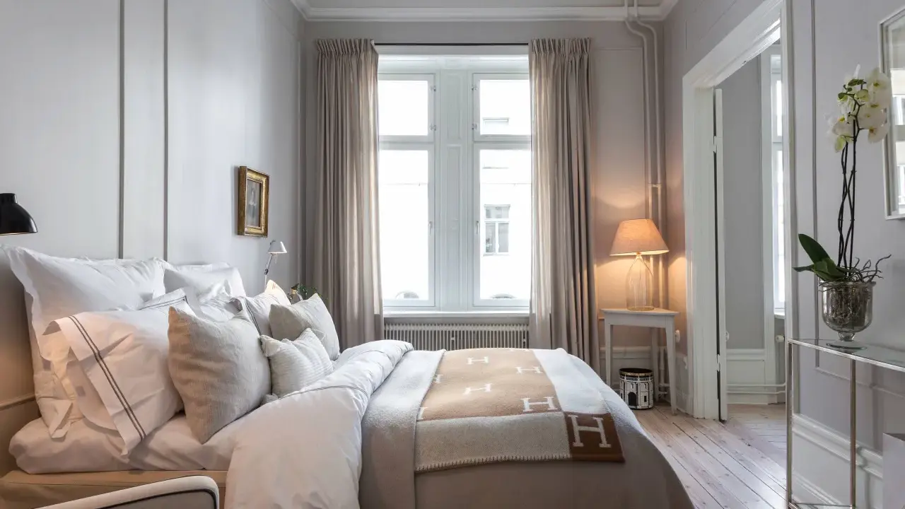 Ett elegant sovrum med en stor säng bäddad med vita lakan och en brun pläd med H-mönster. Vackra möbler till sovrummet som ett litet sidobord med lampa och en orkidé.