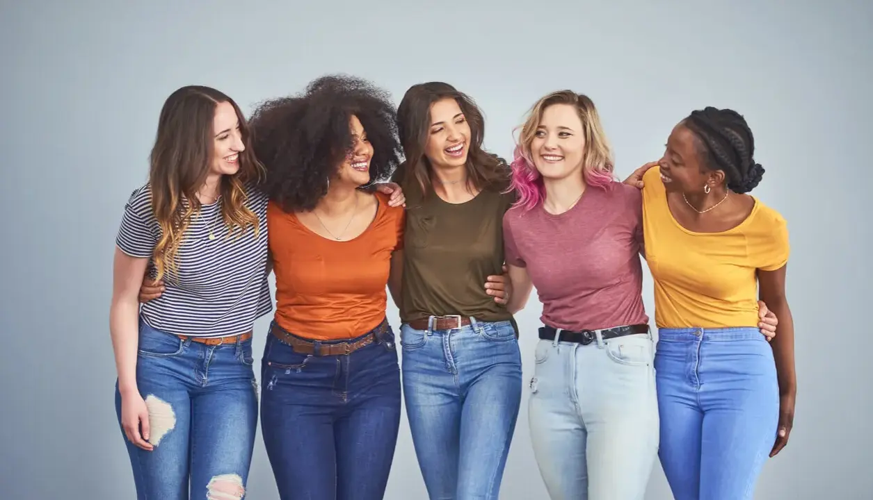 Cinque amiche sorridono, vestite con jeans e magliette colorate, tra cui spiccano tonalità marrone e bianco.