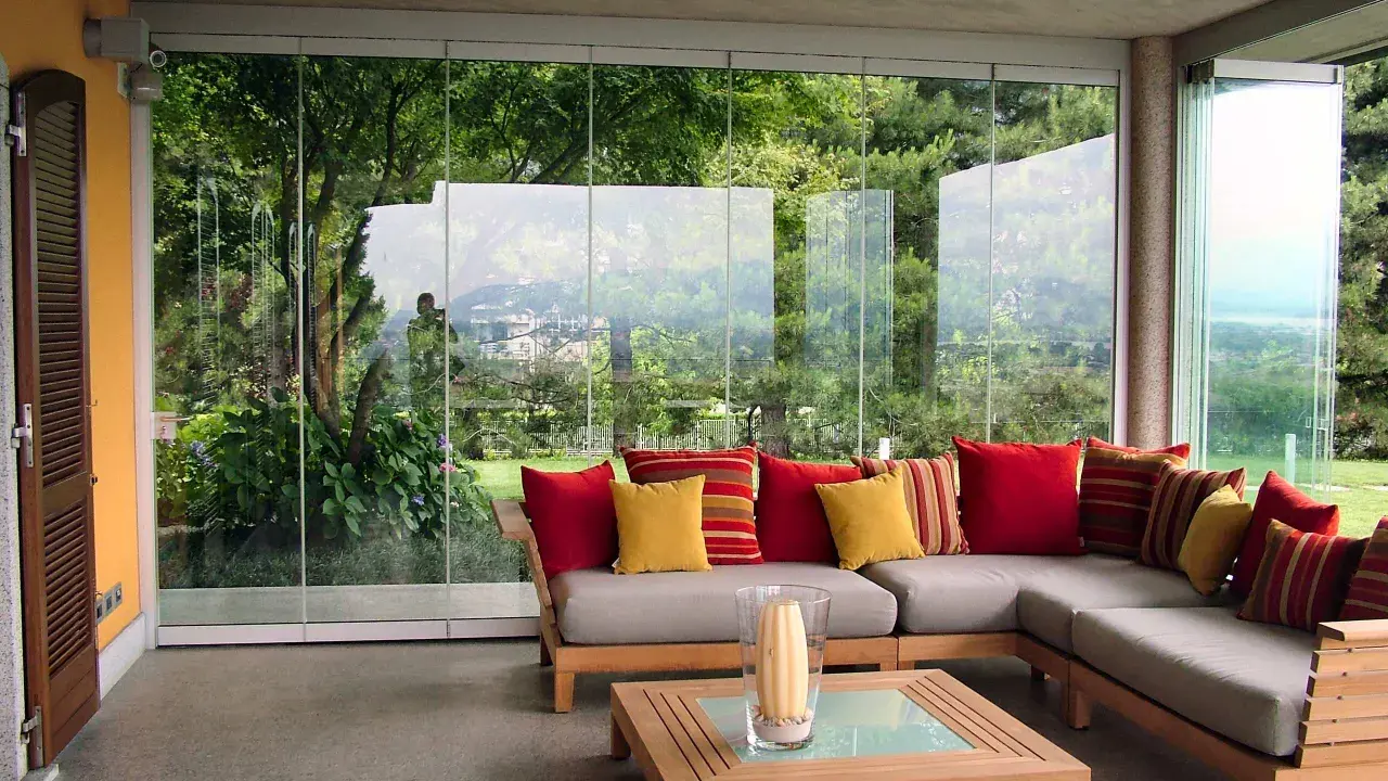 Come arredare una veranda aperta: un divano angolare grigio con cuscini rossi e gialli, un tavolino in legno e una vista sul giardino.
