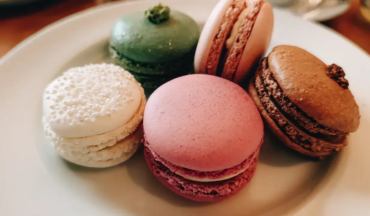 Fem färgglada macarons på en tallrik. En vit med pärlor, en grön, en rosa, en ljusrosa och en brun. Perfekt för ett macarons recept.