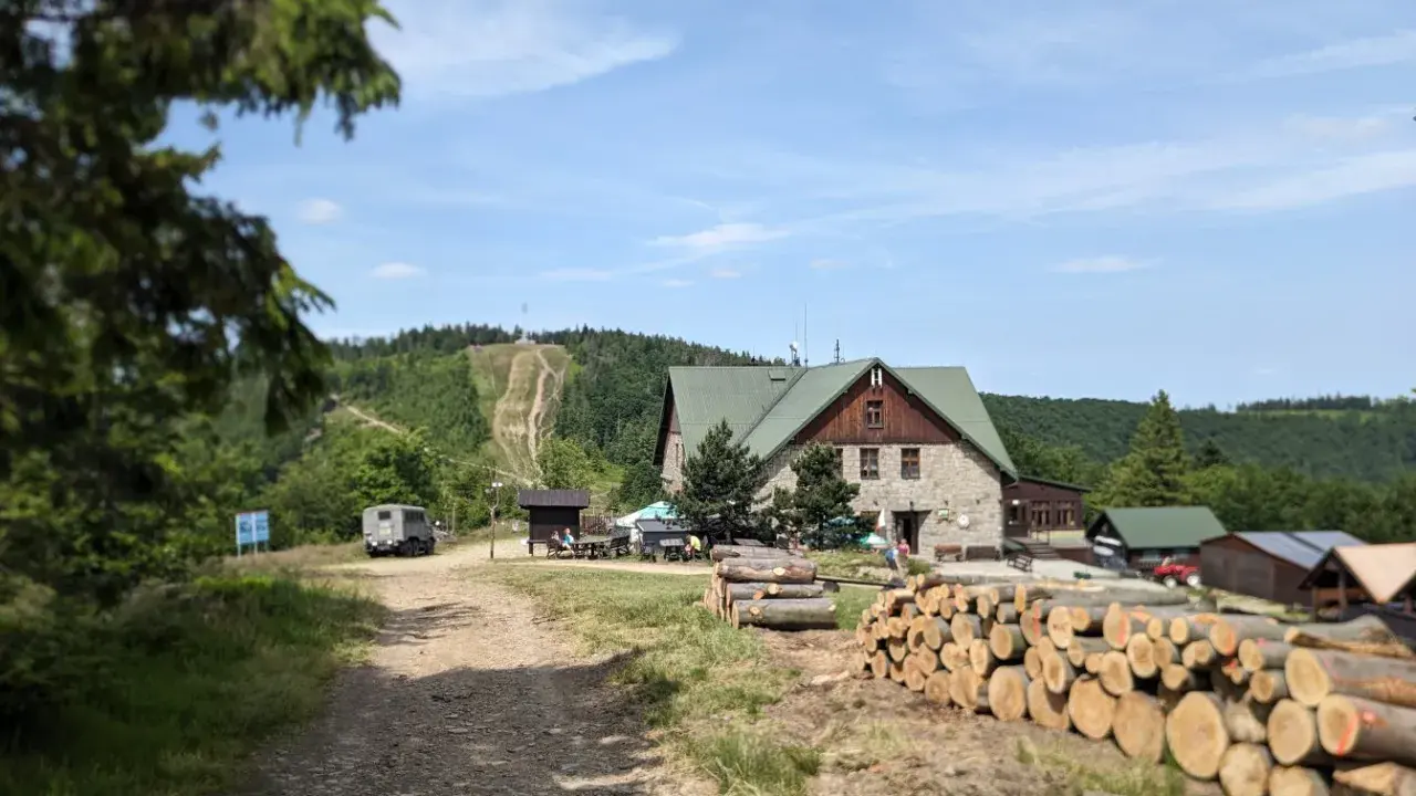 Kamienna chata i stosy drewna przy szlaku na Klimczok. W tle widać górę z wytyczonymi ścieżkami i las.